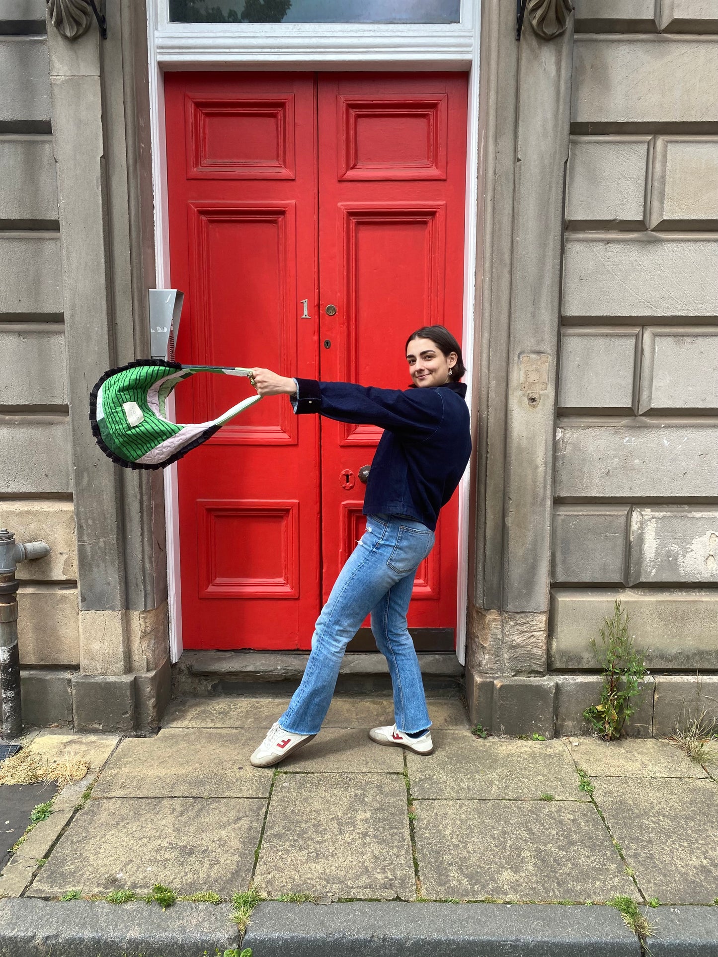 Person holding a green and white Dora Studio bag in front of a red door on a stone building.