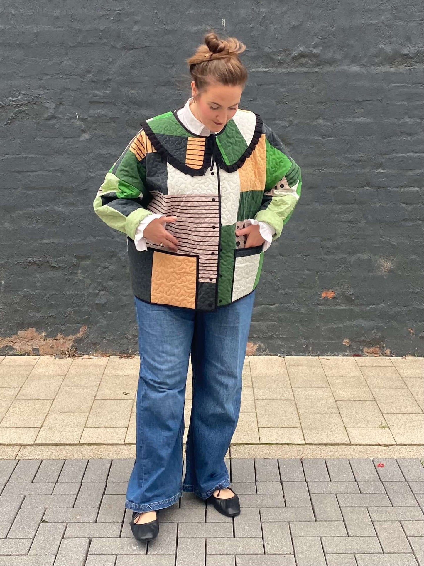 Person wearing a patchwork jacket and jeans standing against a gray wall.