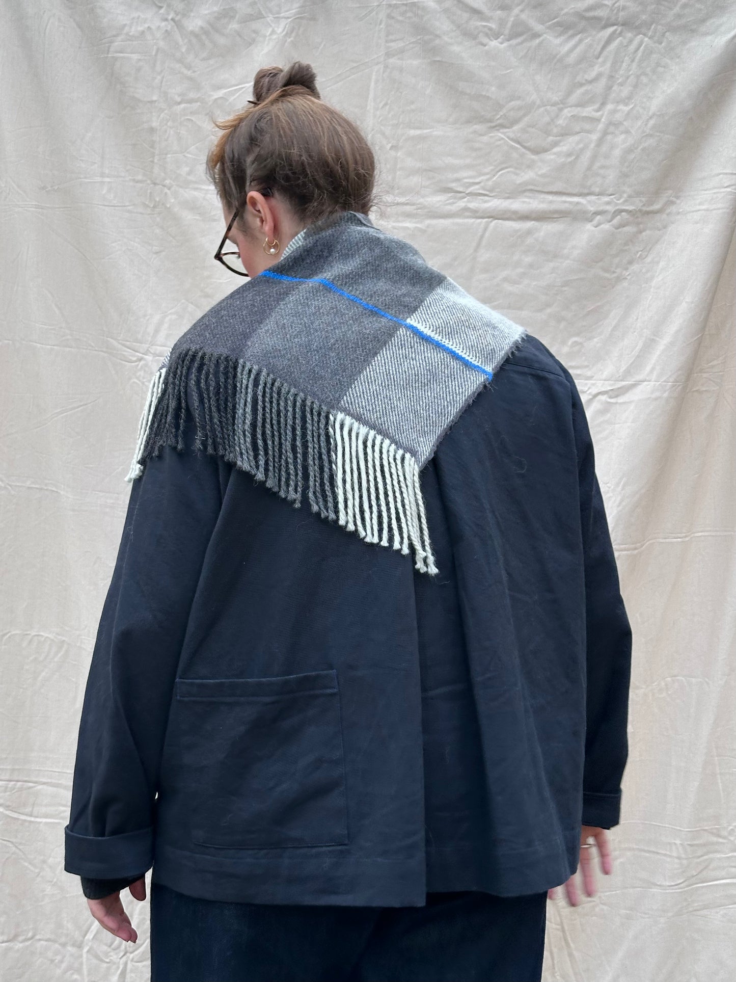 Person wearing a dark jacket with a patterned scarf draped over the shoulders against a plain background