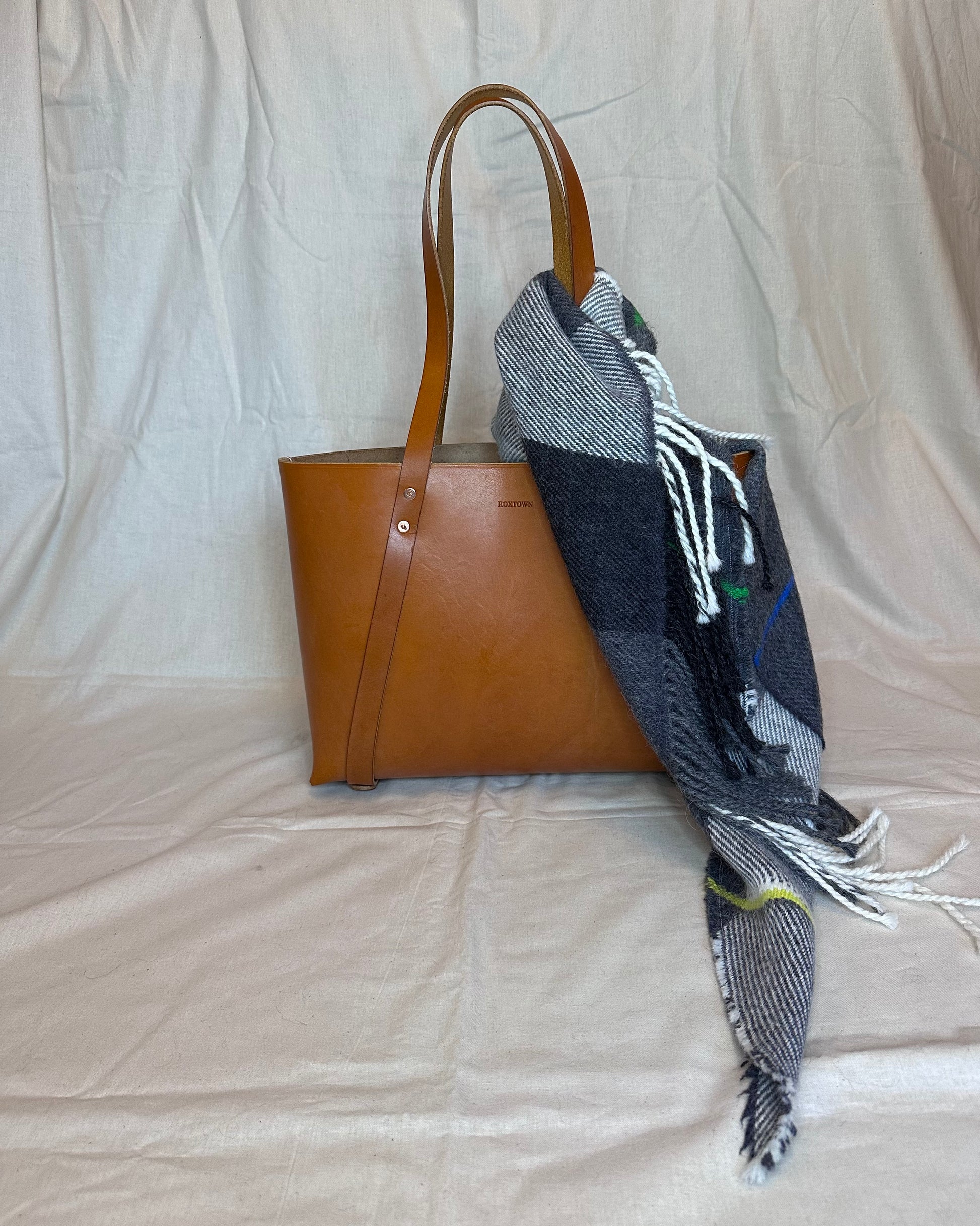 Brown leather tote bag with a plaid scarf draped over it on a plain background